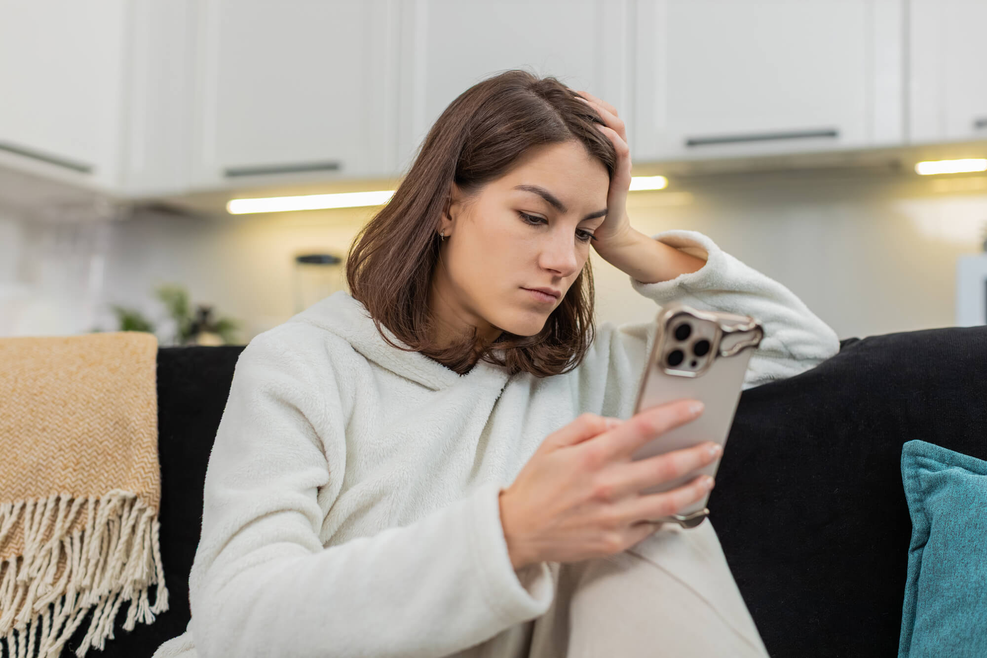 A young woman sitting on a couch at home, looking at her phone with a worried or distracted expression, illustrating restlessness and boredom as recovery triggers